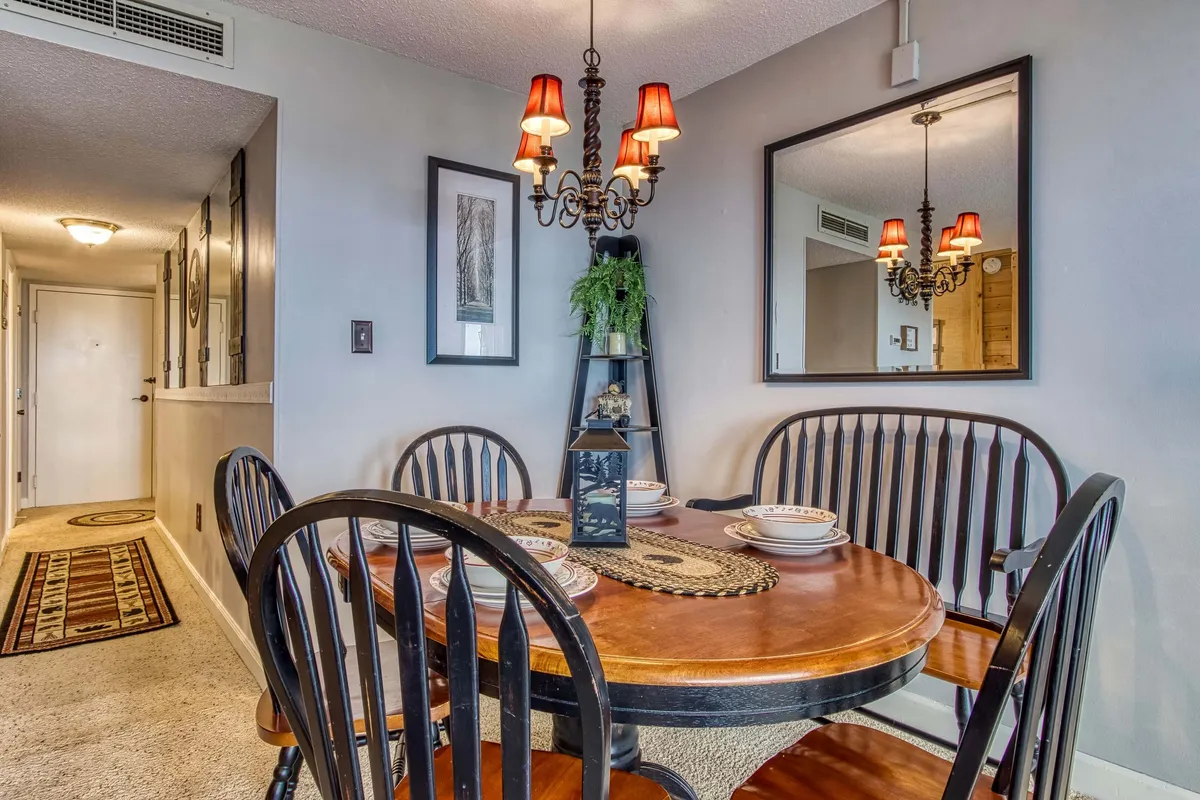 A warm, well-lit dining room centered around a dining table, with a chandelier overhead, wall art, and mirrors adding depth and character. The arrangement is perfect for social gatherings, family dinners, or intimate conversations.