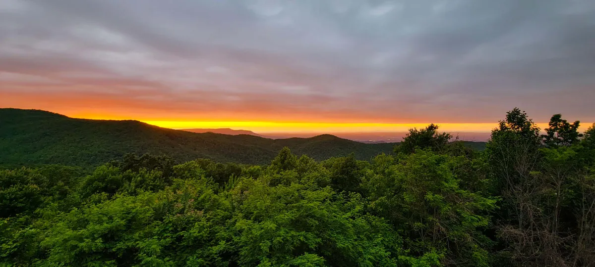 The image showcases a breathtaking view of the Smoky Mountains from a private balcony, with a stunning sunset casting warm hues over the rolling mountain range and lush, dense greenery below. The tranquil atmosphere suggests opportunities for relaxation, wildlife spotting, and unforgettable nature moments.