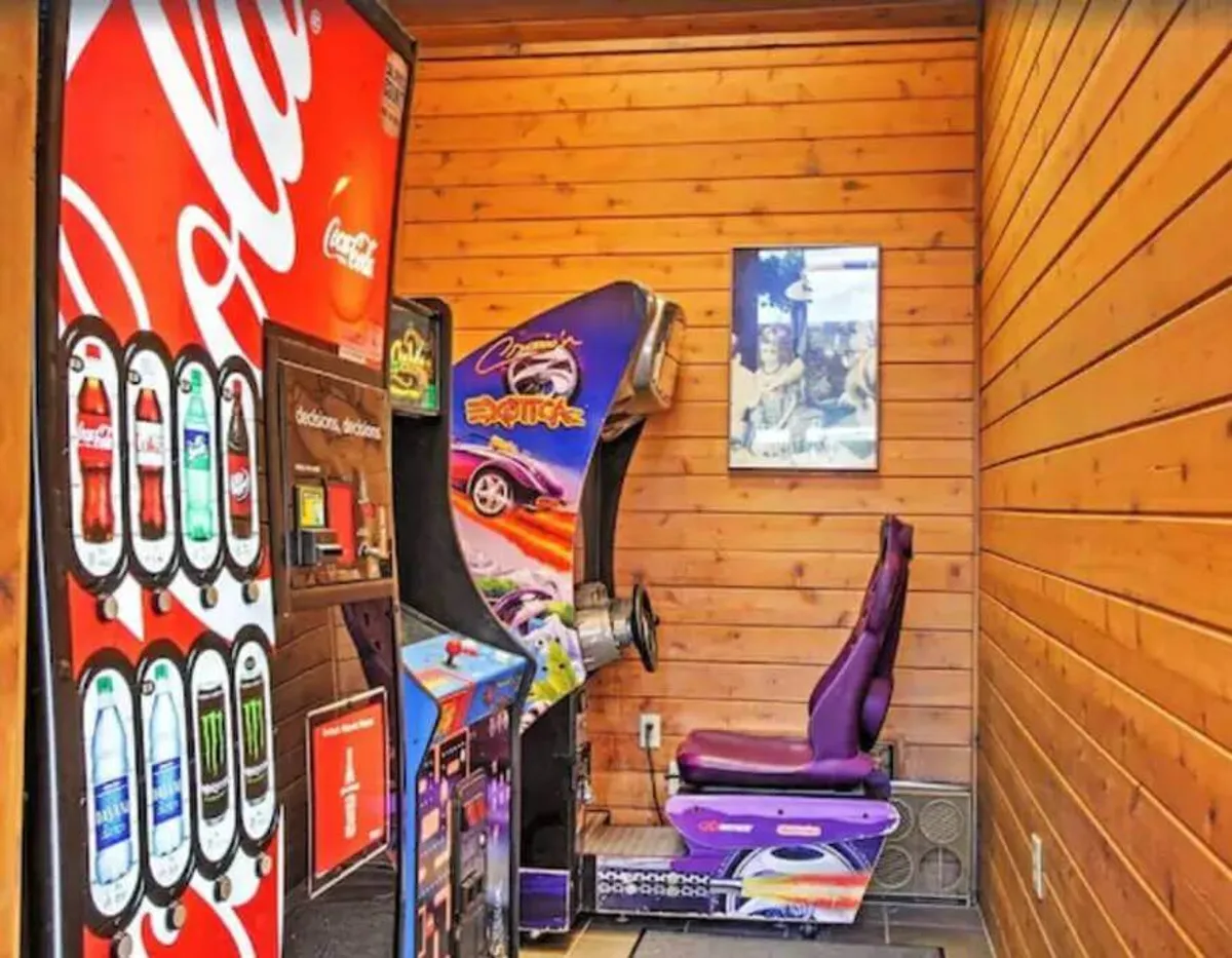 A vibrant arcade room featuring a Coca-Cola vending machine, a retro racing game setup, and a purple racing seat. The space exudes nostalgic fun and is geared toward friendly competition and family entertainment.