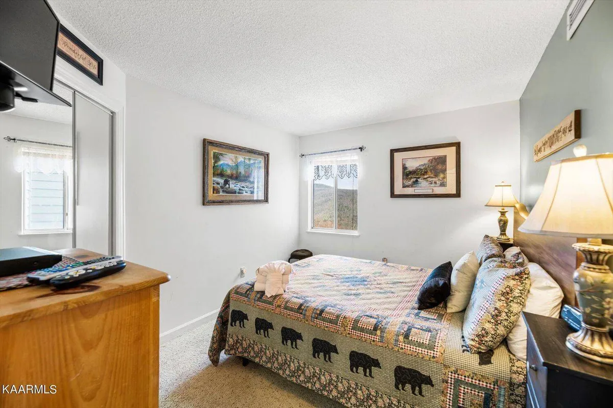 A serene bedroom featuring a bed with a patterned quilt, soft lighting from a bedside lamp, framed artwork, and sunlight filtering through curtained windows. The room has a cozy, welcoming atmosphere ideal for relaxation.