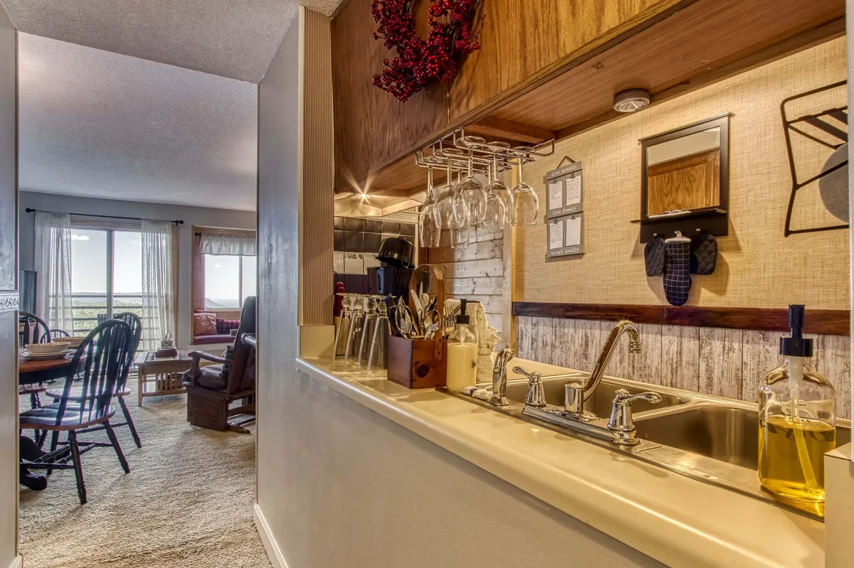 A view from the kitchen sink looking out into the cozy dining area, with hanging glasses above the sink, a welcoming dining table set for a meal, and a glimpse into the adjacent living space. The overall ambiance is warm, inviting, and ideal for both cooking and socializing.