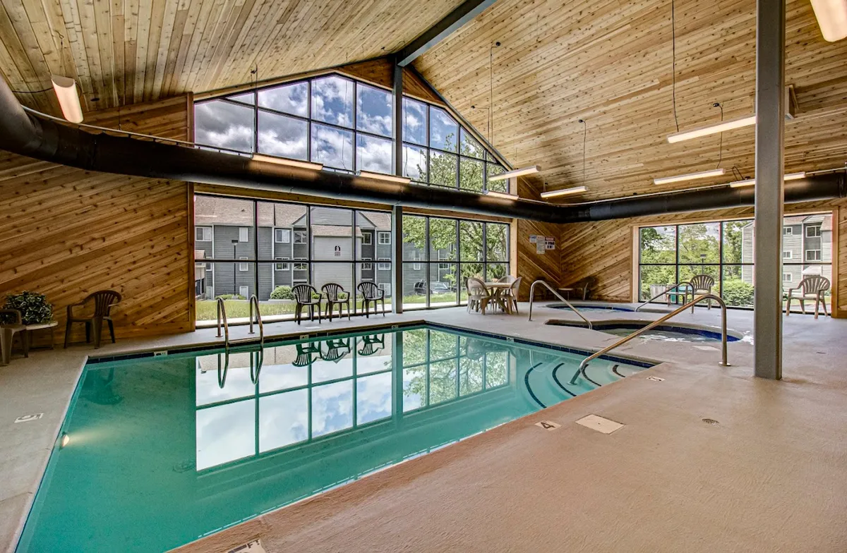 A bright, inviting indoor swimming pool with large windows letting in natural light. The wooden ceiling adds warmth, and there is comfortable seating nearby, creating a sociable, relaxing atmosphere. The space is perfect for swimming laps or lounging by the poolside with friends and family.