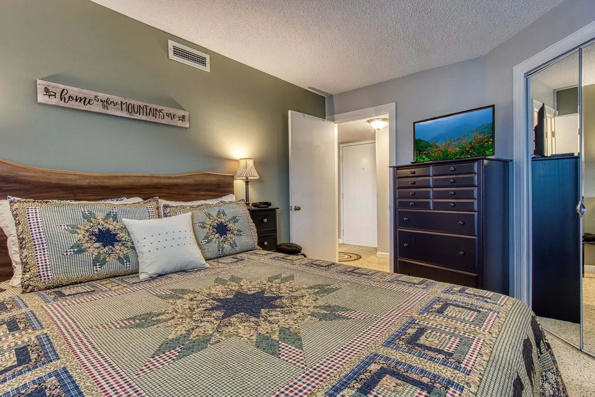 A warmly lit bedroom with a quilted bedspread, nightstand lamp, and a decorative sign. The room feels inviting and is arranged to balance comfort and functionality.