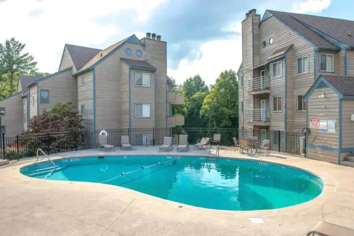 A vibrant outdoor pool scene surrounded by verdant trees and apartment buildings, with families swimming, lounging, and sunbathing. Lounge chairs line the pool, and there's a sense of relaxation and playful energy.