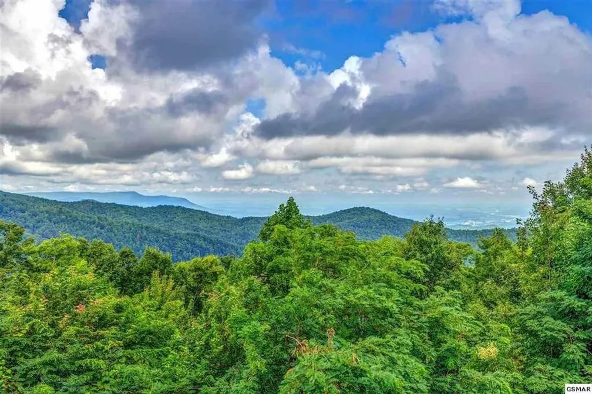 A tranquil view featuring lush green trees in the foreground, a sweeping panorama of distant blue mountains, and dramatic cloud formations above. The scene embodies the pristine natural beauty and vastness of the Great Smoky Mountains, inviting outdoor adventure and peaceful reflection.