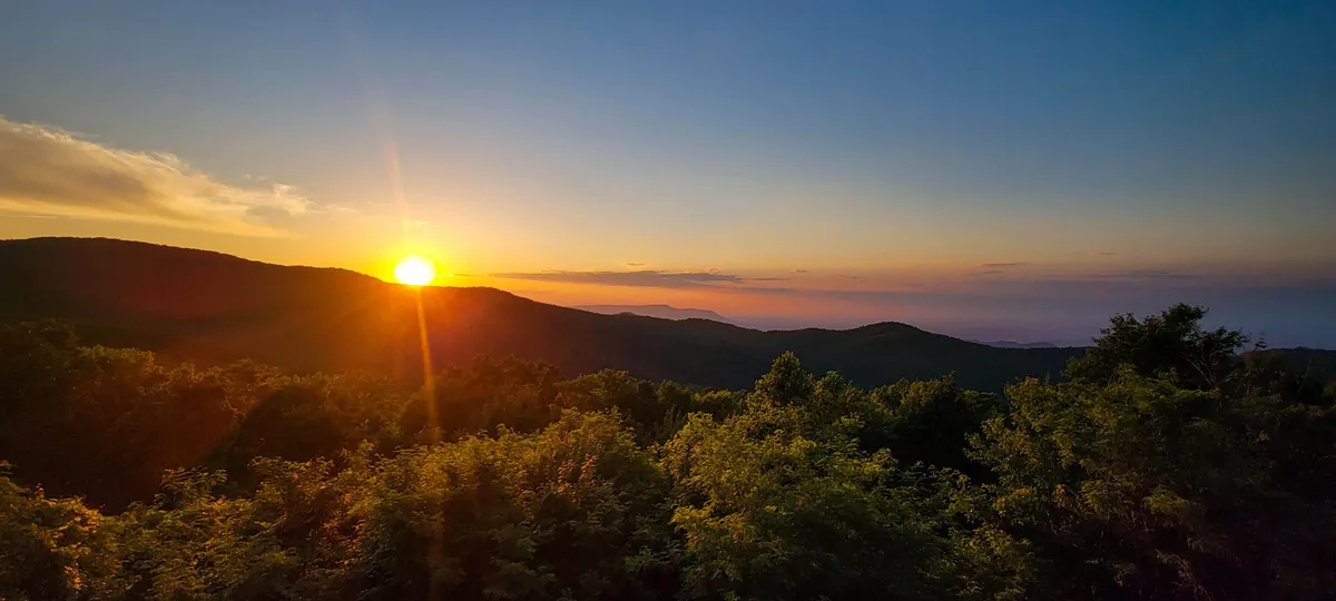 A breathtaking view of the Smoky Mountains at sunset, with layers of blue and green hills stretching into the horizon and lush foliage in the foreground. The sky glows with warm, golden hues, inviting viewers to imagine early mornings or evenings spent soaking in the scenery.