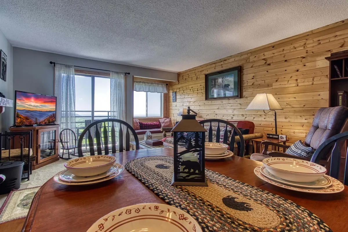 A dining and living area with a set dining table, lantern centerpiece, and a comfortable seating area positioned beside expansive windows. The windows capture natural light and offer glimpses of the outdoor scenery, seamlessly blending indoor relaxation with the beauty of the mountains.